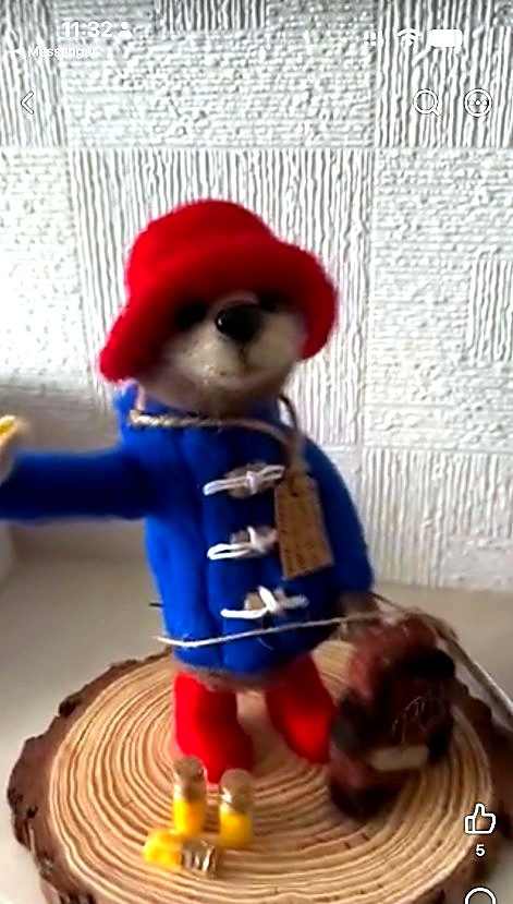 Needle-felted bear in a blue duffle coat and red hat, standing on a wood slice with tiny marmalade jars